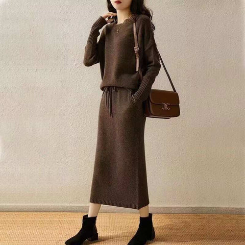 Woman wearing coffee-colored knitted loose slimming top with long sleeves and matching mid-length skirt two-piece set with a brown shoulder bag and black ankle boots against a plain beige wall