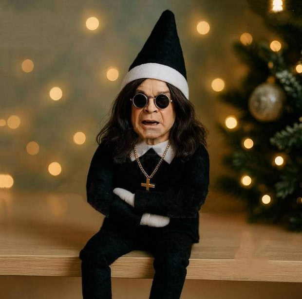 Christmas Oz Doll resin elf decoration in black outfit with hat and sunglasses on wooden surface with blurred Christmas lights background